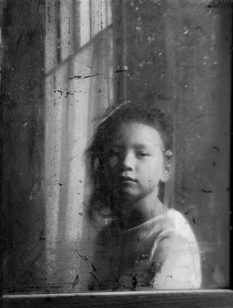 Black and white portrait of a young Asian child taken through a window with distressed glass.